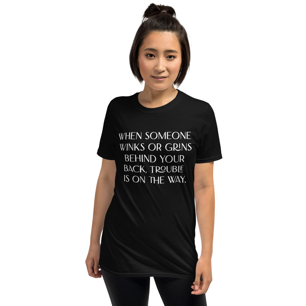Unisex soft-style black T-shirt with WHEN SOMEONE WINKS OR GRINS BEHIND YUR BACK, TROUBLE IS ON THE WAY in White letters