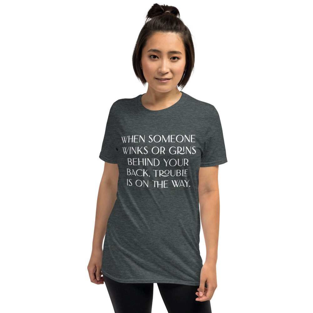 Unisex soft-style dark heather T-shirt with WHEN SOMEONE WINKS OR GRINS BEHIND YOUR BACK, TROUBLE IS ON THE WAY in White letters